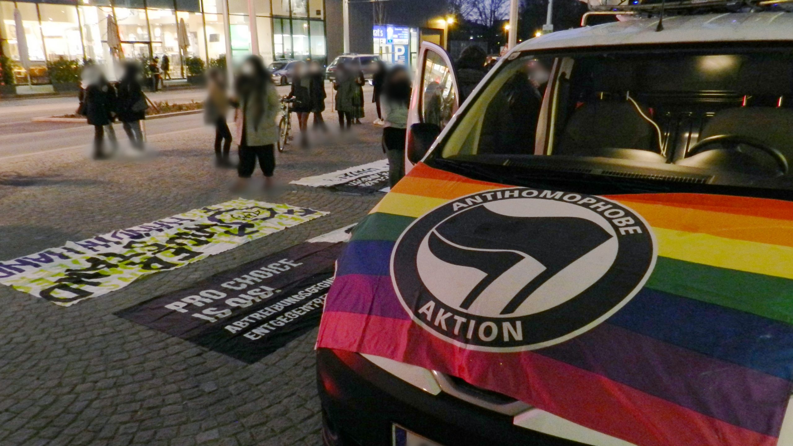 Foto des Demo-Autos. Auf der Motorhaube ist eine Regenbogenfahne mit Antifa-Symbol befestigt ("Antihomophobe Aktion"). Im Hintergrund sieht man mehrere feministische Transparente.